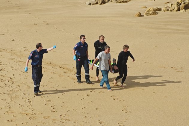 16895828 - paramedic in uniform points direction to group of surfers and another paramedic who carry injured surfer in a stretcher and walk on sand at bells beach, rocks in background, victoria, australia on december 15